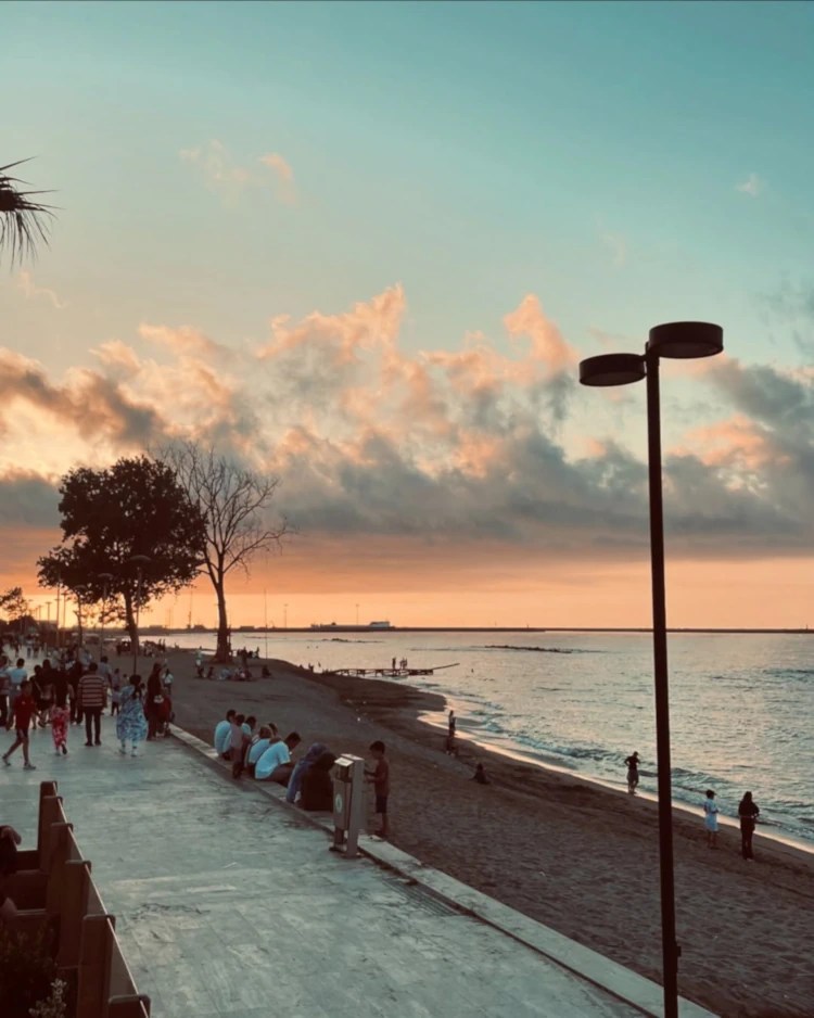A crowded coastline, with people walking on a boardwalk and sitting on the sand as the sun sets over the Black Sea.