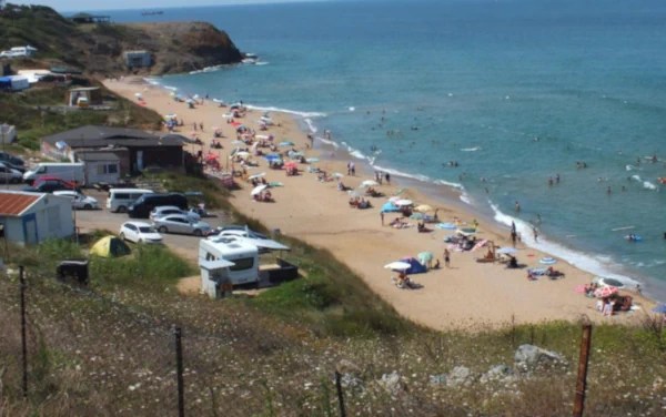 A crowded beach with a sandy shore and calm blue water. Sunbathers and swimmers fill the area, with small buildings and hills in the background.
