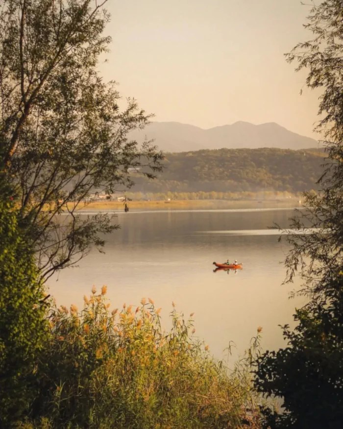 A still Sapanca Lake with distant hills, framed by foliage. A single orange kayak floats centrally.