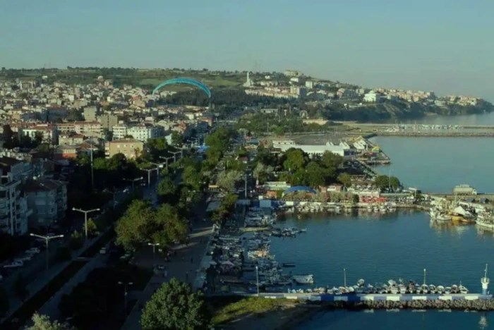 A wide aerial shot of the Süleymaniye district of Tekirdağ, Turkey, showing a coastal town with buildings built along a hillside leading down to a harbor filled with boats.