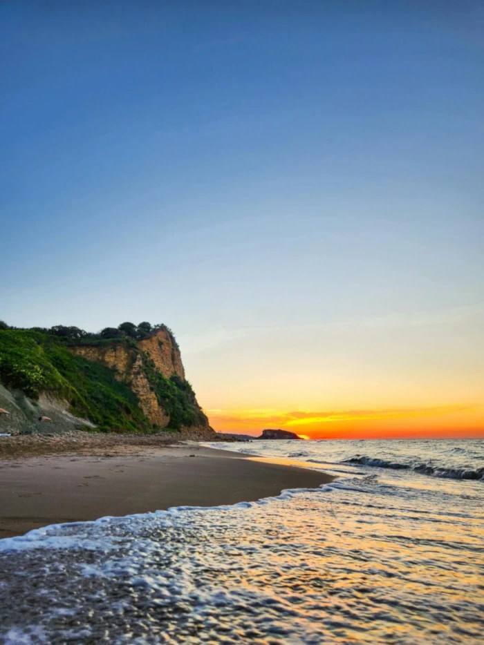A sunset view from a sandy beach, with a prominent cliff on the left and a calm sea on the right under an orange and blue gradient sky.