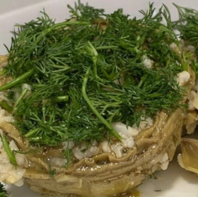 Close-up of a single artichoke heart stuffed with a mixture of rice and other ingredients, topped with a large sprig of fresh dill. The artichoke sits on a white plate.