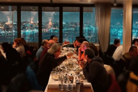 A lively Istanbul tavern with large windows overlooking the city at night, filled with people dining, drinking, and enjoying conversations.