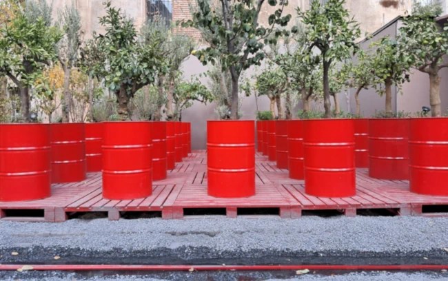 A close-up of Khalil Rabah's "Red Navigapparate" installation, showing multiple bright red oil barrels placed on a raised platform of wooden pallets, each barrel containing a small green tree, set against a weathered, beige brick wall backdrop.