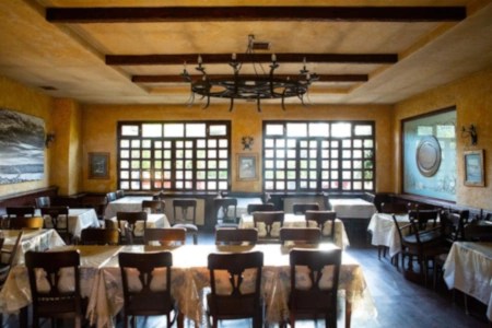 An empty traditional tavern with rustic yellow walls, wooden beams, and neatly arranged tables covered in white cloths.
