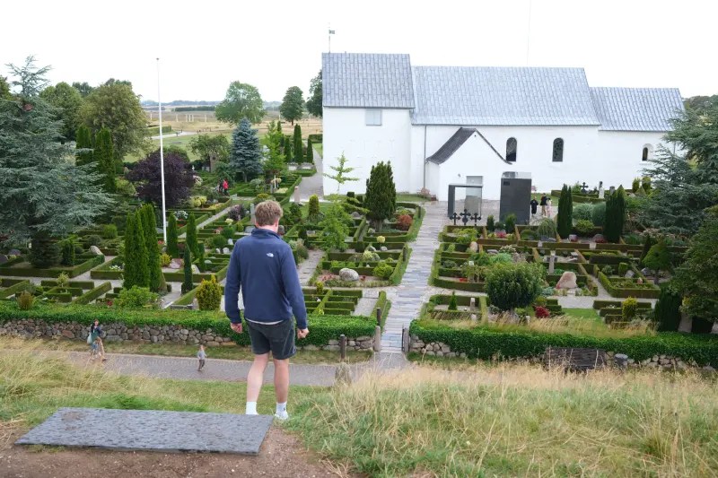 Man loopt van trapje van grafheuvel bij Jelling Kirke. Op de voorgrond zie je een modern kerkhof. In één van de grafheuvels zou Gorm de Oude hebben gelegen, vader van Harald Blauwtand.