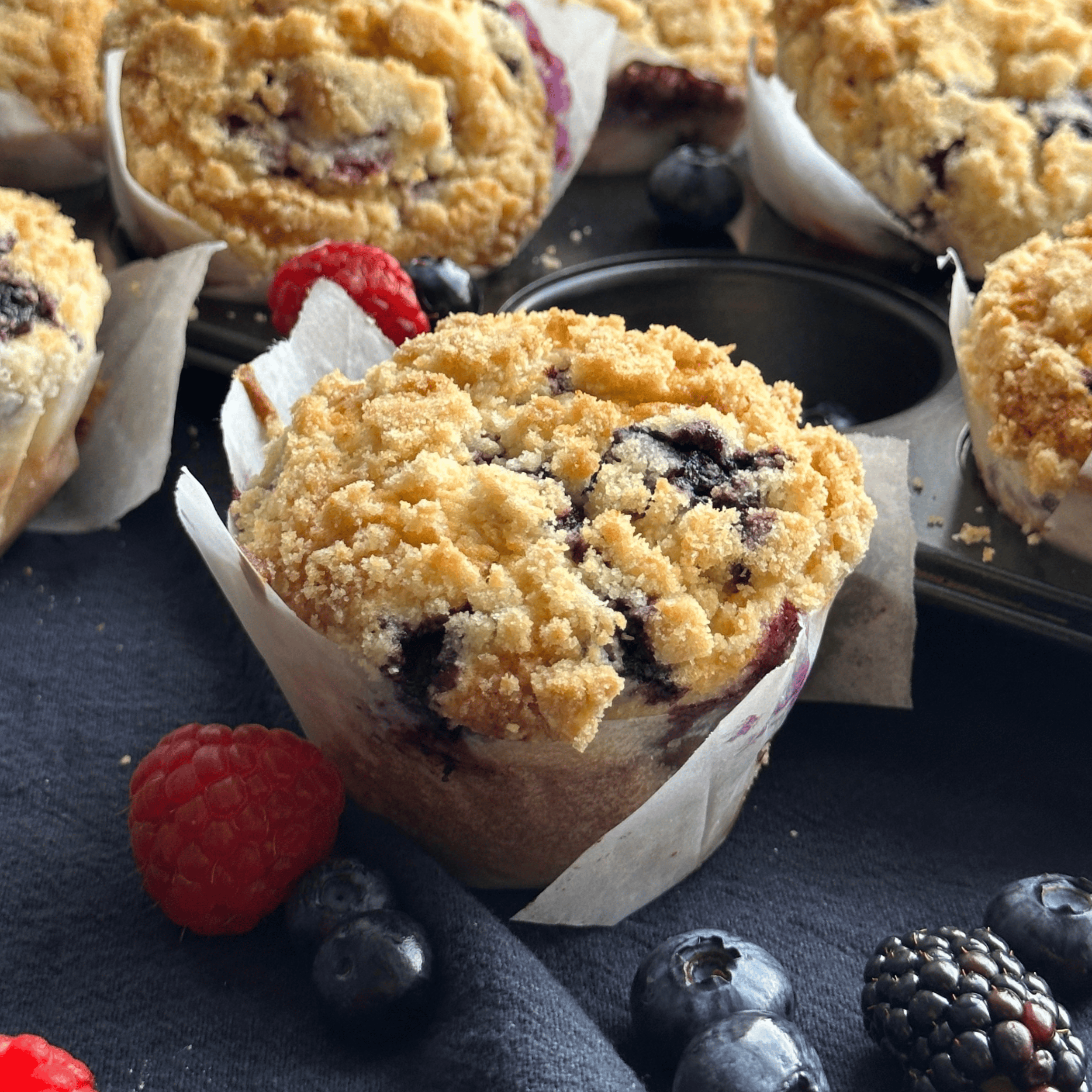 a muffin in the middle of the image with raspberries, blackberries, and blueberries around. The background has more muffins laying in its tray.
