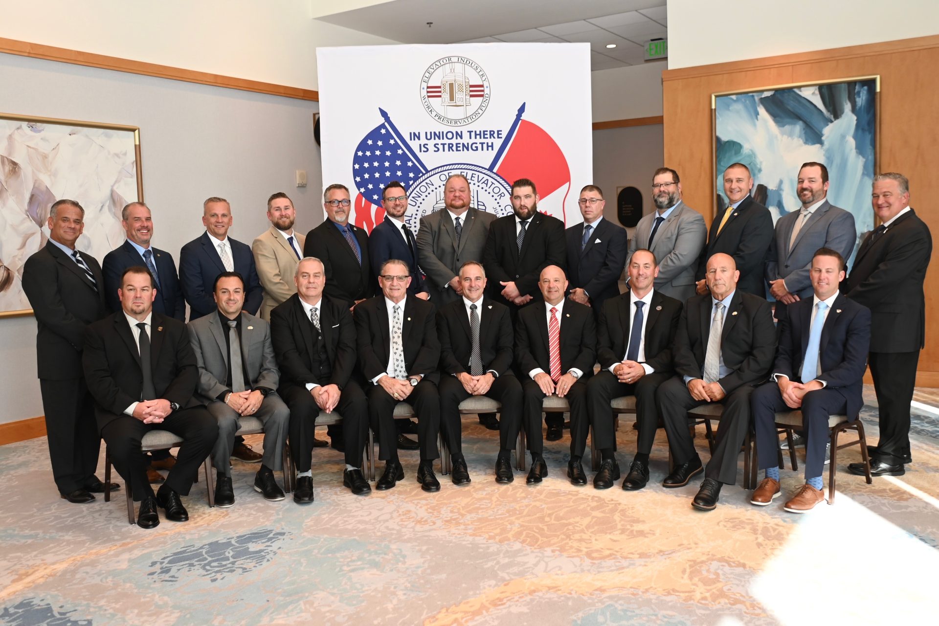 IUEC - A group of 21 men in suits pose for a formal photo in front of a banner with U.S. and Canadian flags and the phrase IN UNION THERE IS STRENGTH. Some are seated, others stand behind them in two rows indoors. International Union of Elevator Constructors
