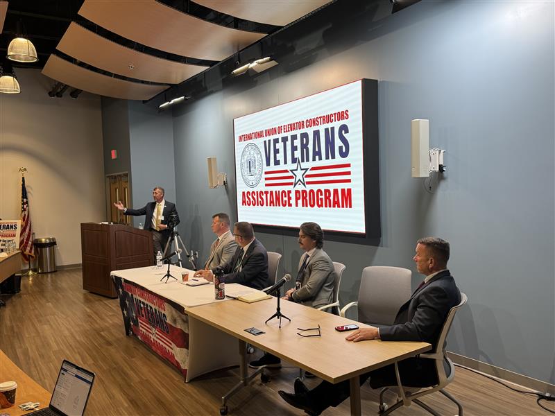 IUEC - A man stands at a podium speaking to an audience, while four men from the International Union of Elevator Constructors sit at a panel table beside him. A large screen behind displays Veterans Assistance Program in a conference room setting. International Union of Elevator Constructors