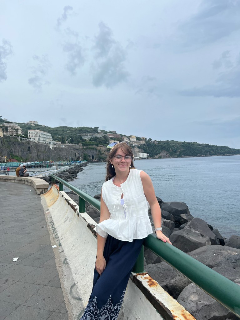 Photo of Jamisyn standing in front of the sea leaning against a green railing on a path