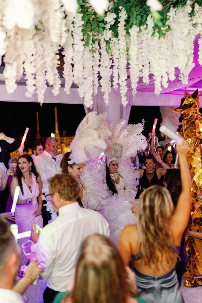 Wedding guests dancing with white feathered Hora Loca performers and a golden mirror stilt walker under hanging floral décor.