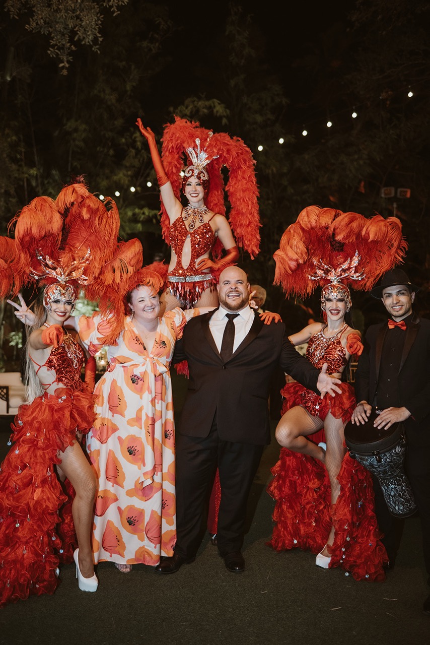 Wedding guests posing with Vegas showgirls in red feather costumes and drummer at Villa Woodbine celebration.