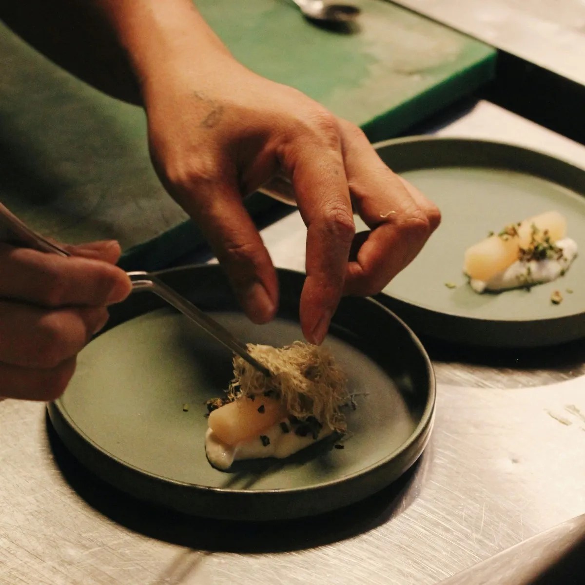 Hands placing a garnish with tweezers on a green ceramic plate in a dim kitchen