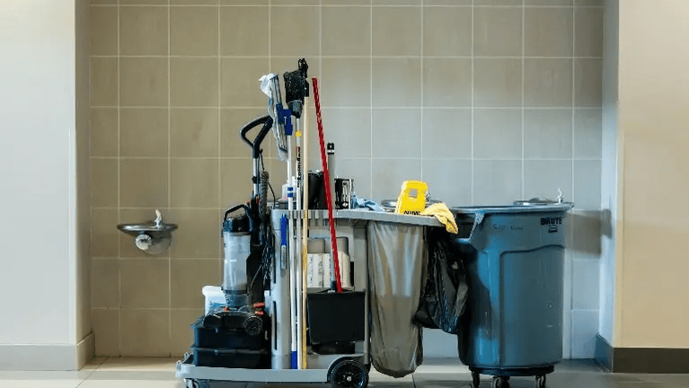 A janitorial cart with cleaning supplies, mops, and a trash bin is positioned against a tiled wall, illustrating how to choose a janitorial service for your business.