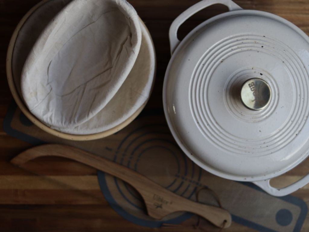 a dutch oven, banneton sourdough bread proofing baskets, breqad knife and a silicone baking sling on a wooden cutting board