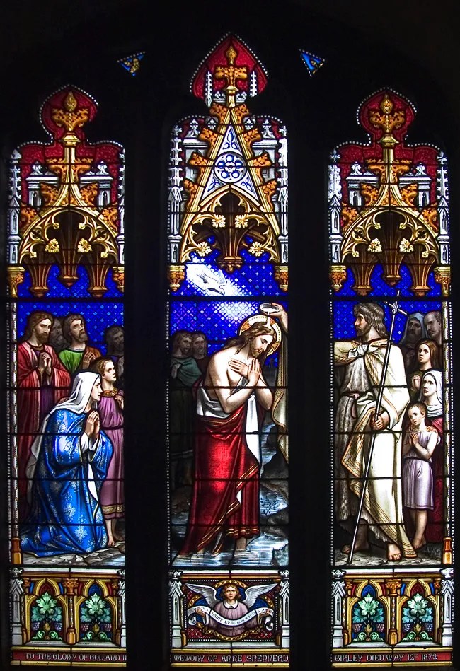 A three light stained glass window showing the Baptism of Christ with the dove of the Holy Spirit descending upon the head of Christ.