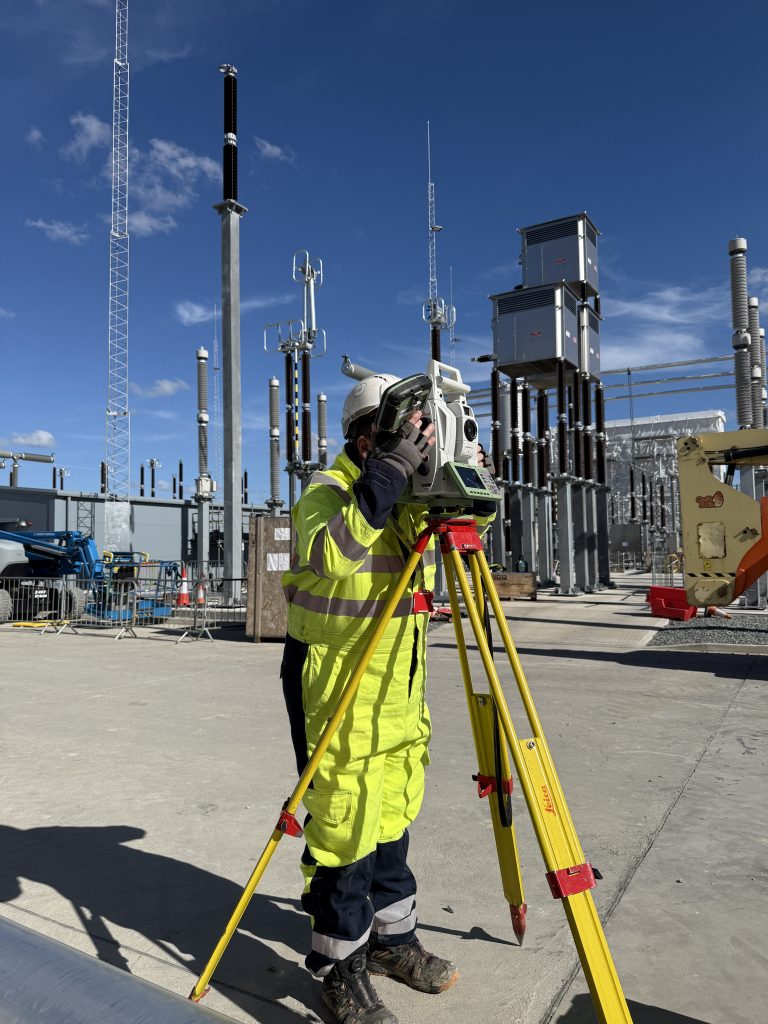 surveyor looking through total station with electrical infrastructure in the background
