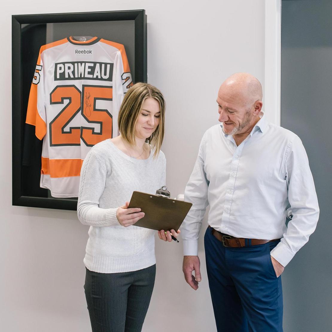 Woman and man conferring over a clipboard with a Primeau Number 25 autographed orange and white jersey hanging behind them.