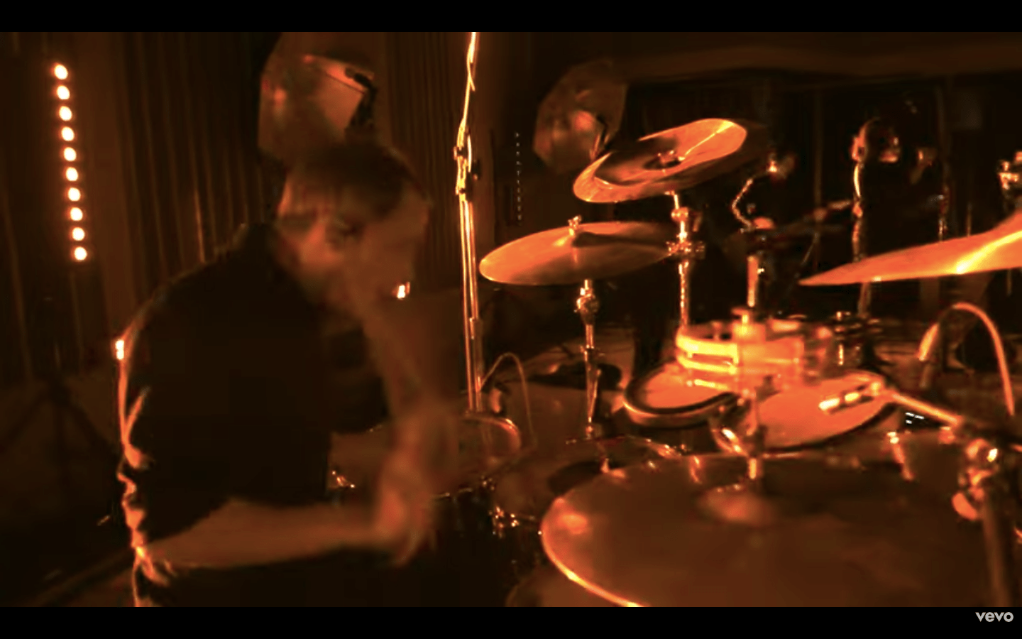Rock drummer performing intense live set in studio, cymbals crashing under warm orange stage lights, band in background