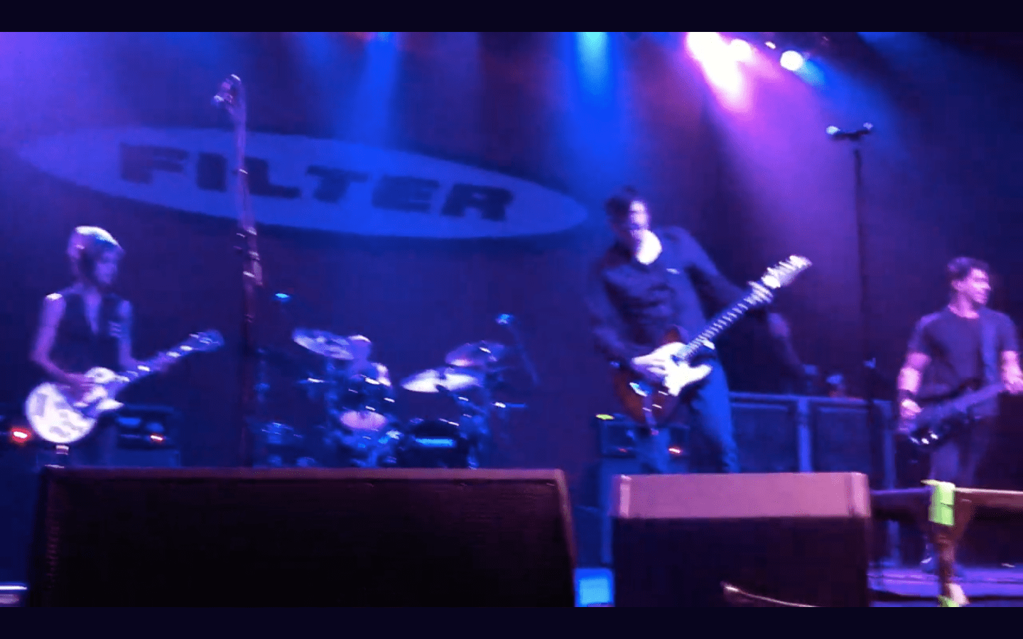 Alternative rock band performing under 'FILTER' banner; guitarist, drummer, bassist onstage with purple concert lighting.
