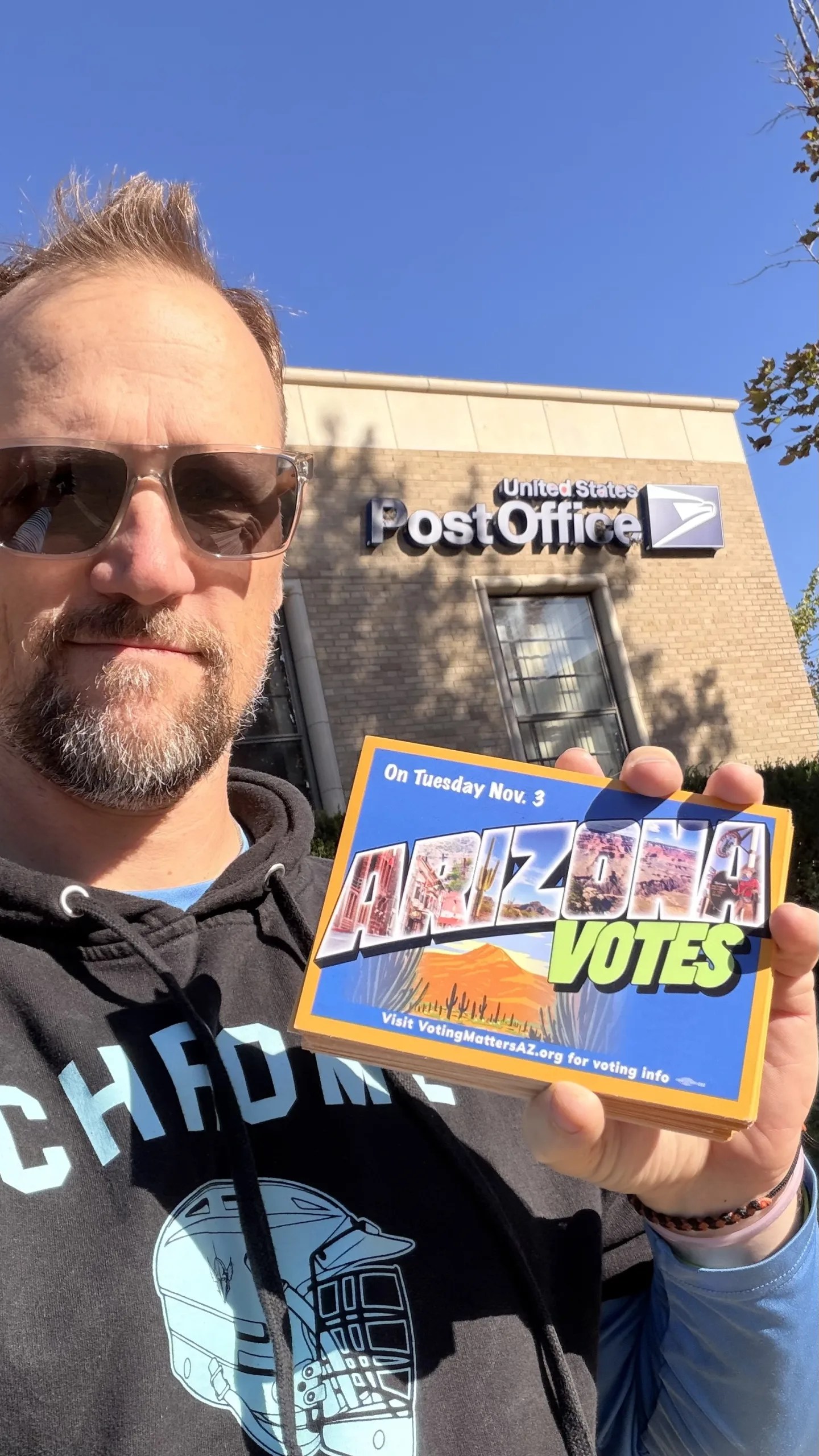 Selfie of JP holding up “Arizona Votes” post cards in front of a post office