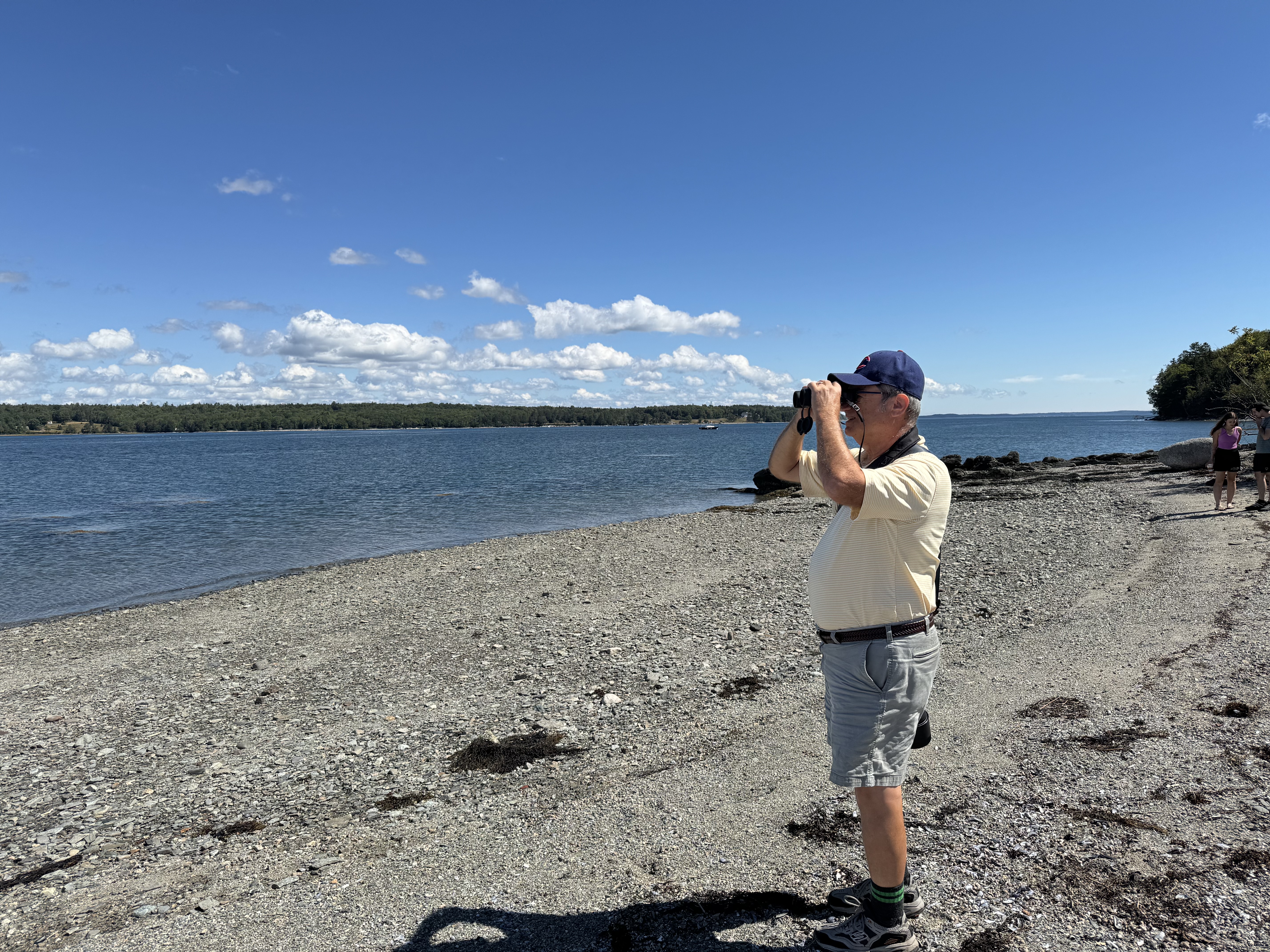 Man in baseball caps using binoculars to look out on the water while standing on beach.