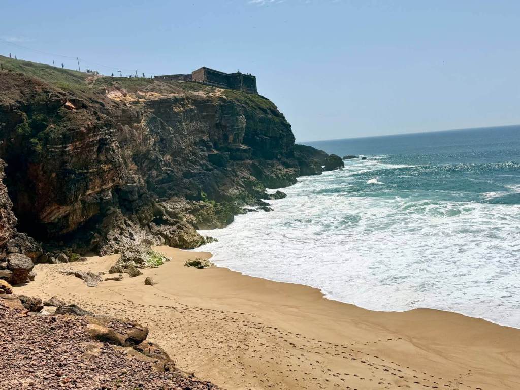 A sandy beach with ocean waves crashing against a rocky cliff topped by an old stone building under a clear blue sky.