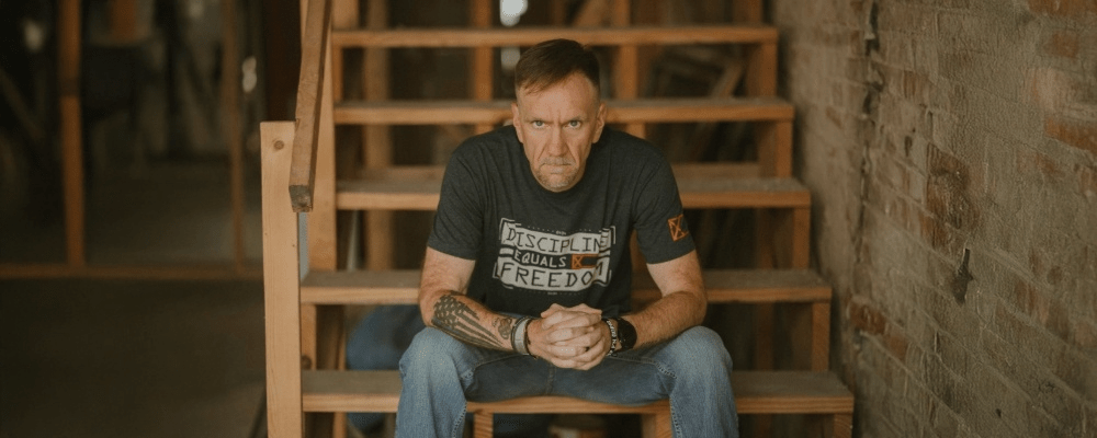 Jim Lunsford is sitting on wooden stairs in a dark setting, wearing a 'Discipline Equals Freedom' shirt, staring intensely with hands clasped.