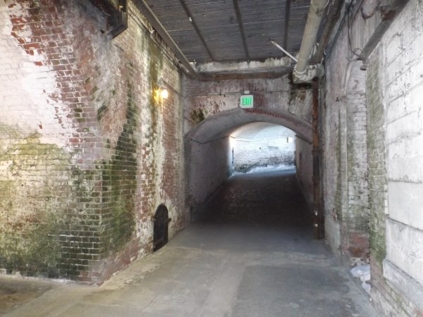 Auto-generated description: A dimly lit brick tunnel with an exit sign leads to a brighter area in the distance.