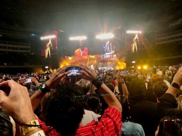 Auto-generated description: A large crowd at a concert captures the moment with their phones as performers play on a brightly lit stage.