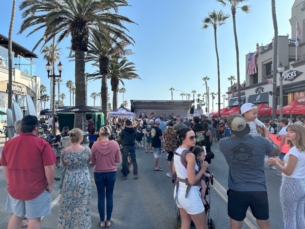 Auto-generated description: A crowd of people is gathered on a street lined with palm trees, engaging in an outdoor event with booths and a stage.