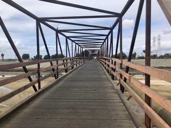 Auto-generated description: A long wooden bridge with a geometric metal framework spans across a dry landscape under a cloudy sky.