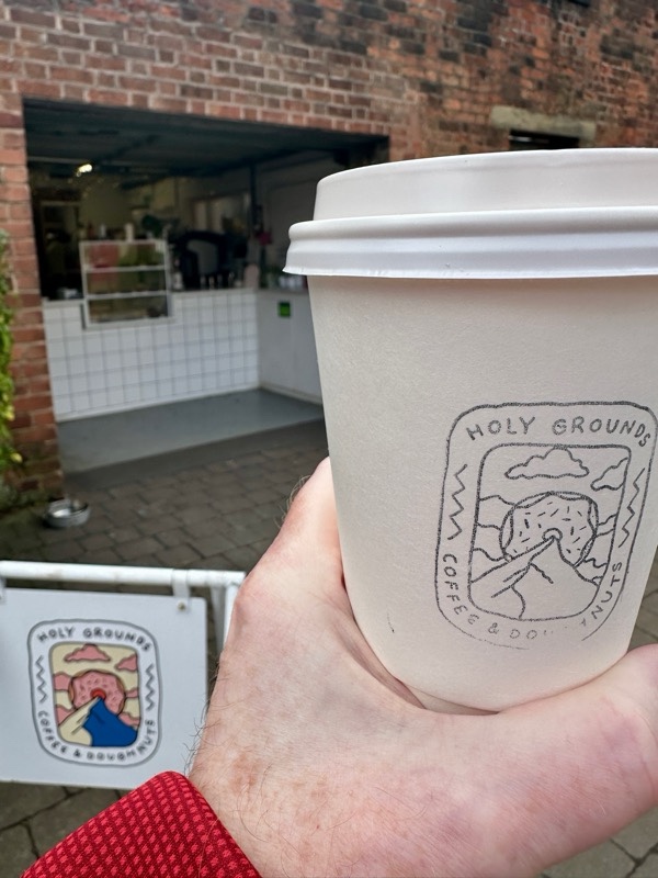A disposable coffee cup in a hand in front of Holy Grounds Coffee and Doughnut in Preston