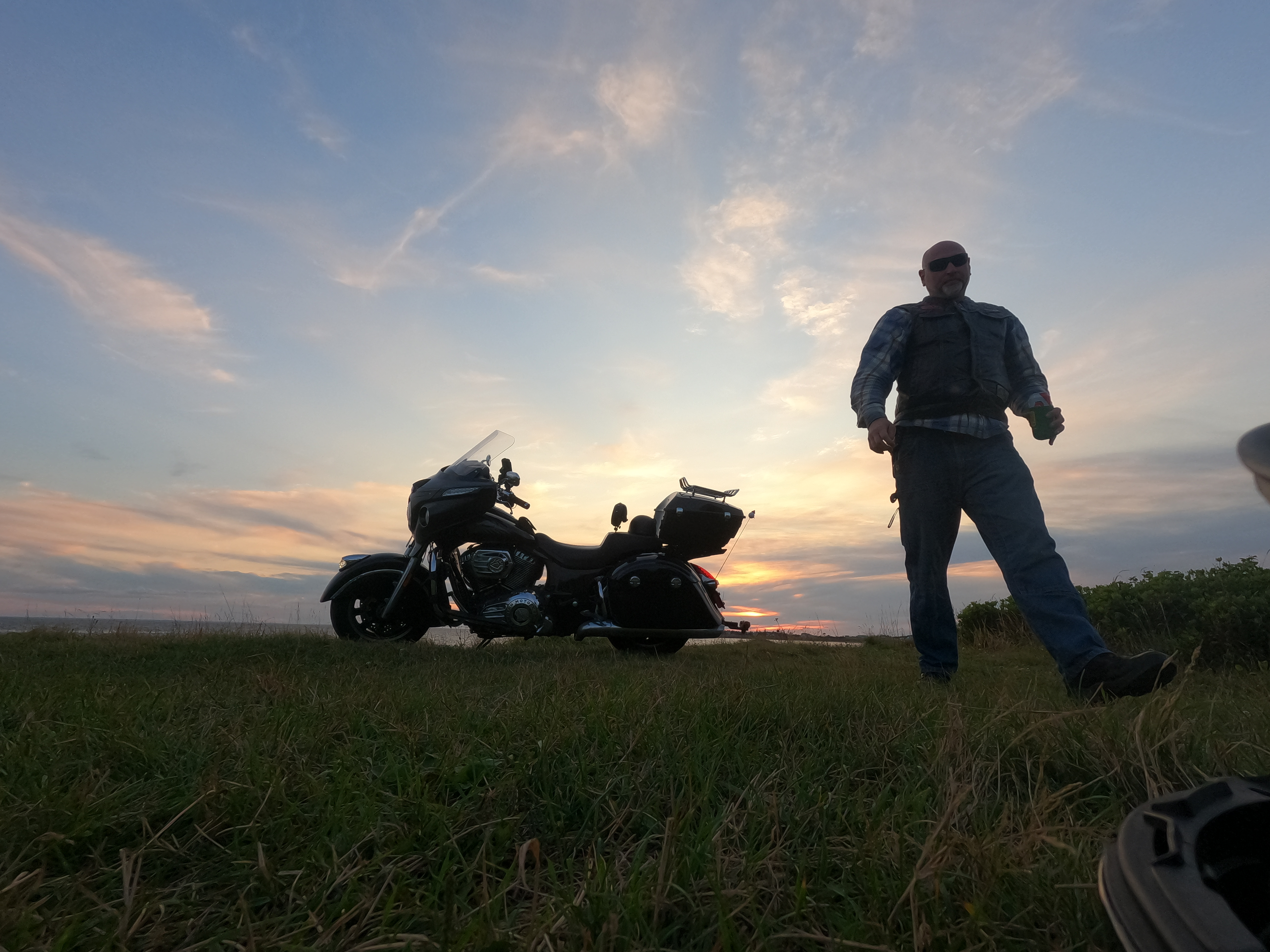 golden clouds lit by a setting sun. the camera sits at ground level, looking out to the silhouette of a motorcycle on the kickstand, a man is also in silhouette walking to the camera