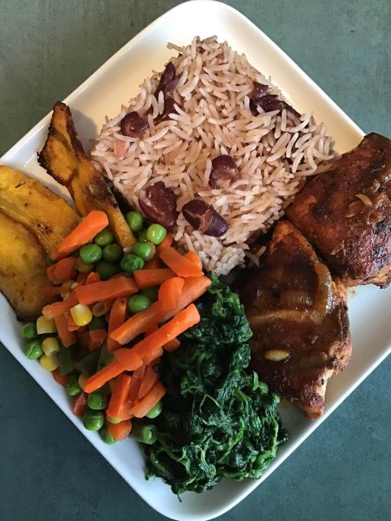A serving dish of jamaican rice and peas, chicken, fried plantains, spinach and mixed vegetables.
