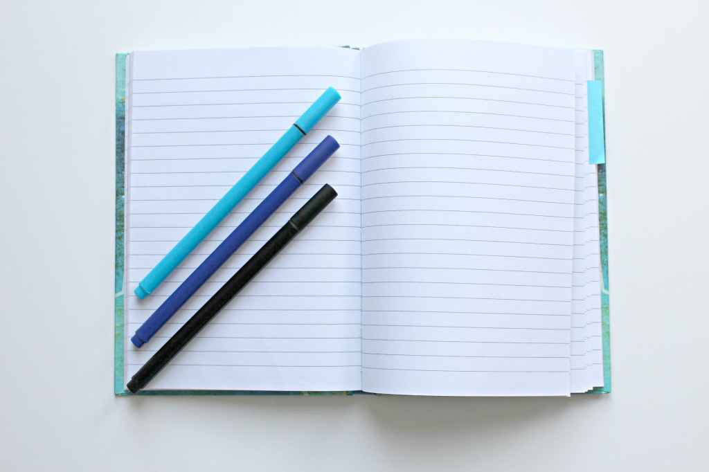 Foto de um caderno aberto com folhas em branco, e em cima dele três lápis em diagonal: o de cima azul-claro, o do meio azul-marinho e o de baixo preto, representando o propósito de estudar e aprender coisas novas. Fim da descrição.