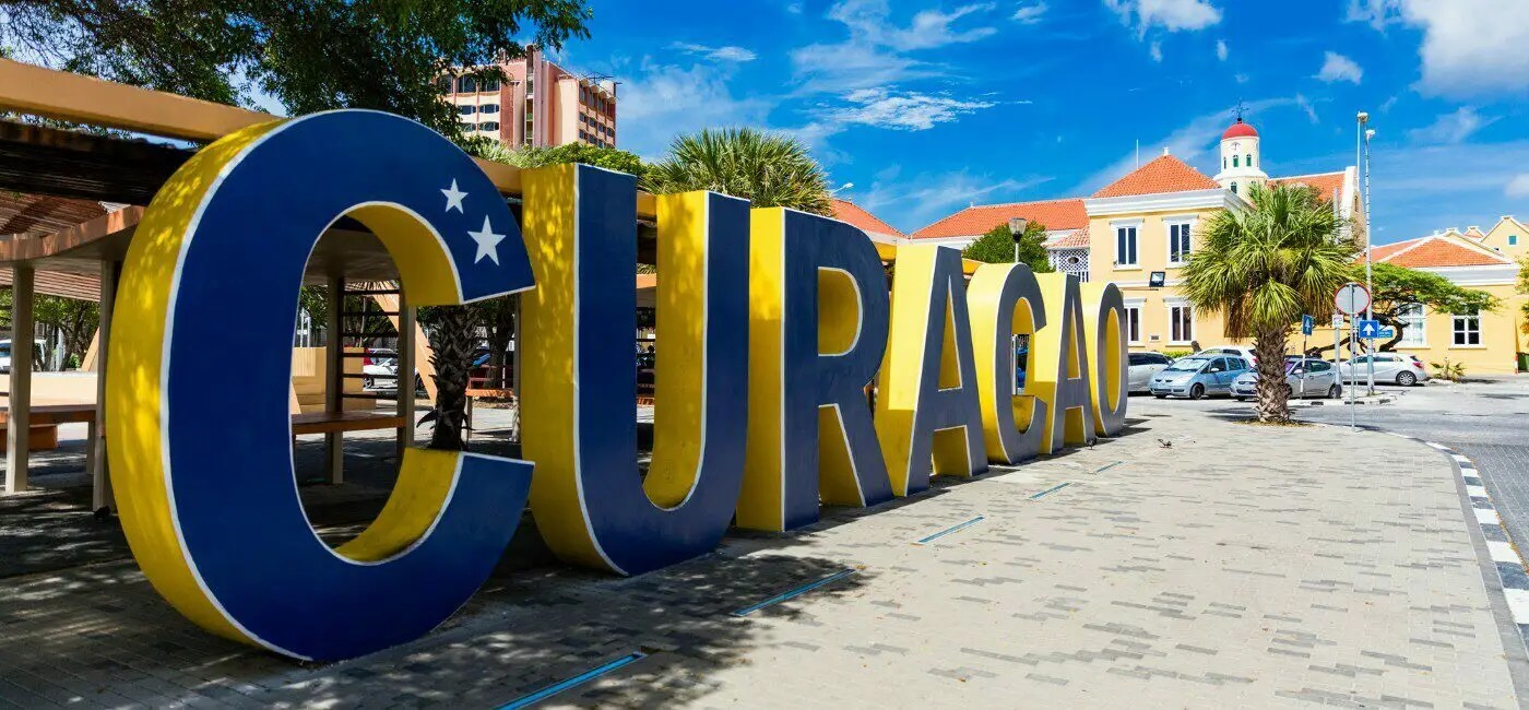 Large yellow and blue CURA&Ccedil;AO sign in Willemstad with colonial buildings and palm trees in the background