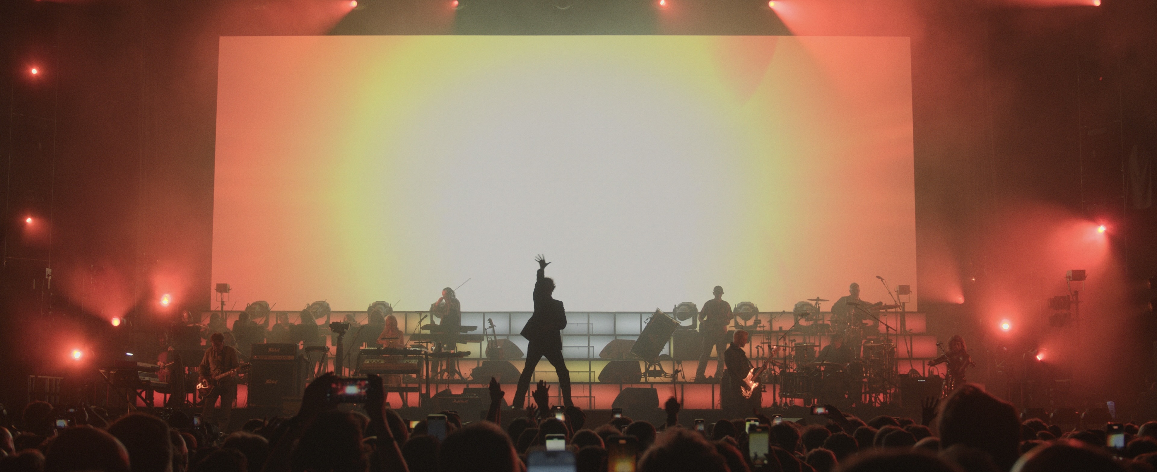 Pulp performing onstage at the O2 in front of a bright orange 'sunburst' backdrop. Jarvis Cocker is in silhouette and has thrust his hand straight up in the air.