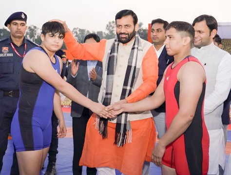 एक तस्वीर जिसमें दो पहलवान, एक नीले और दूसरा लाल कुश्ती परिधान में, आपस में हाथ मिला रहे हैं। बीच में एक प्रतिष्ठित व्यक्ति, नारंगी कुर्ता और शॉल पहने, एक पहलवान के सिर पर आशीर्वाद भरा हाथ रखे हुए हैं। पृष्ठभूमि में सुरक्षाकर्मी और अन्य लोग दिखाई दे रहे हैं।