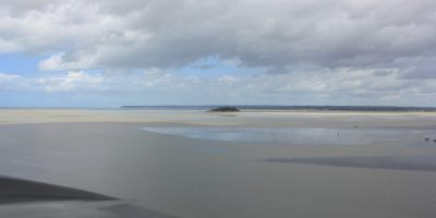 Hier ist ein weitläufiger Strand zu sehen, das Meer hat sich zurückgezogen, und unter einem bewölkten Himmel ist knapp über dem Horizont ein blauer Streifen zu sehen.