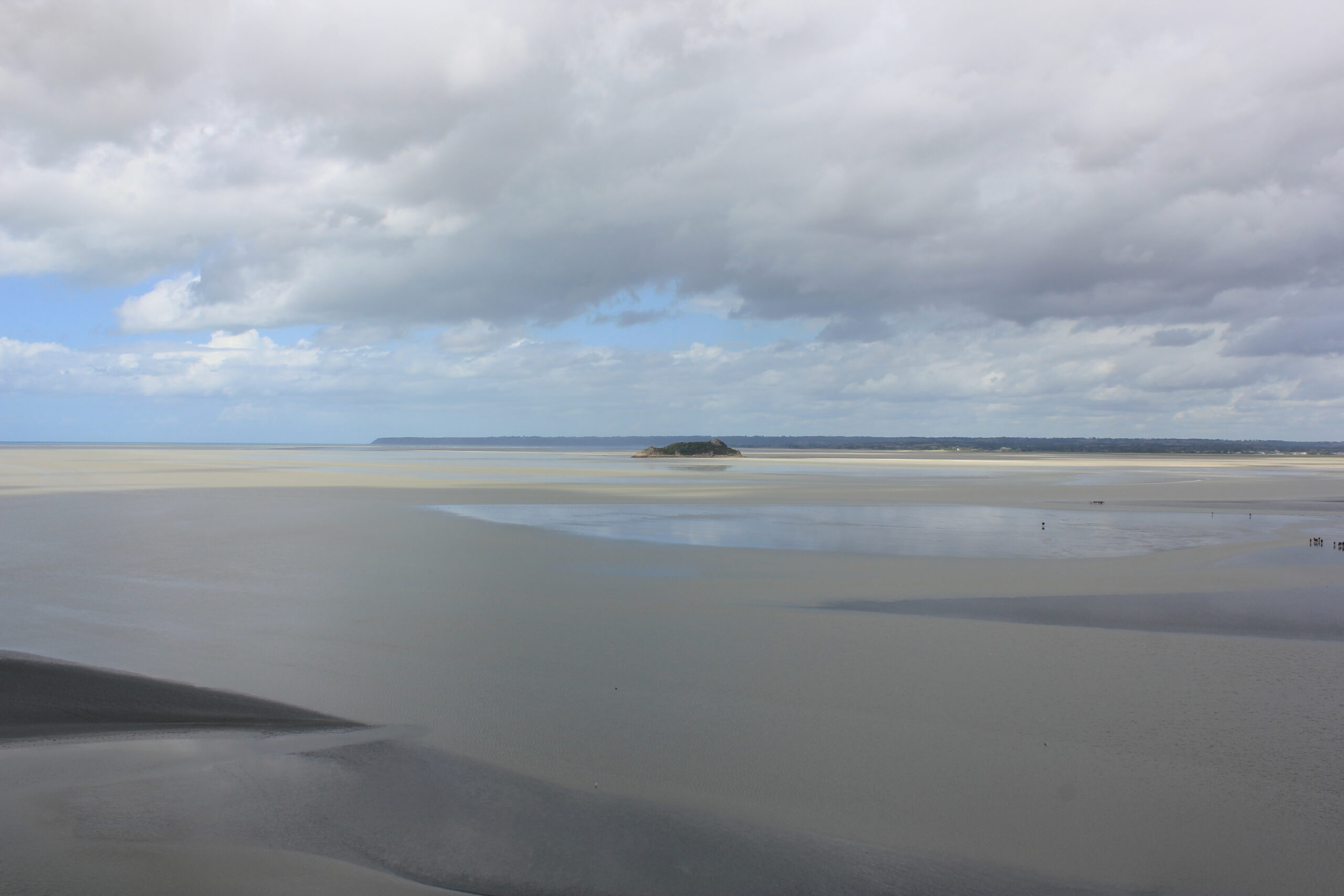 Hier ist ein weitläufiger Strand zu sehen, das Meer hat sich zurückgezogen, und unter einem bewölkten Himmel ist knapp über dem Horizont ein blauer Streifen zu sehen.