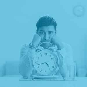 Photo of a man sitting at a table looking frustrated. He has his face leaning on his fists. A large clock sits right in front of him in the center of the photo.