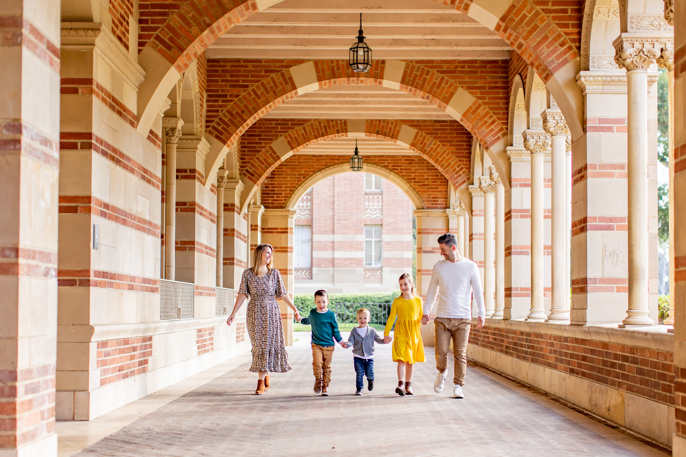 A mom, dad, and three small kids walk together holding hands and laughing along a brick hallway at UCLA during a fall mini session