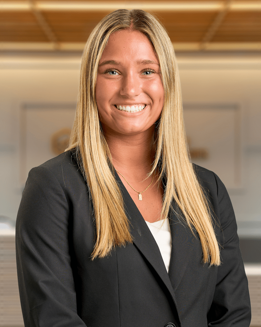Ally, a woman with long blonde hair smiling in a black blazer with a gold necklace, professional headshot with a warm lit office background.
