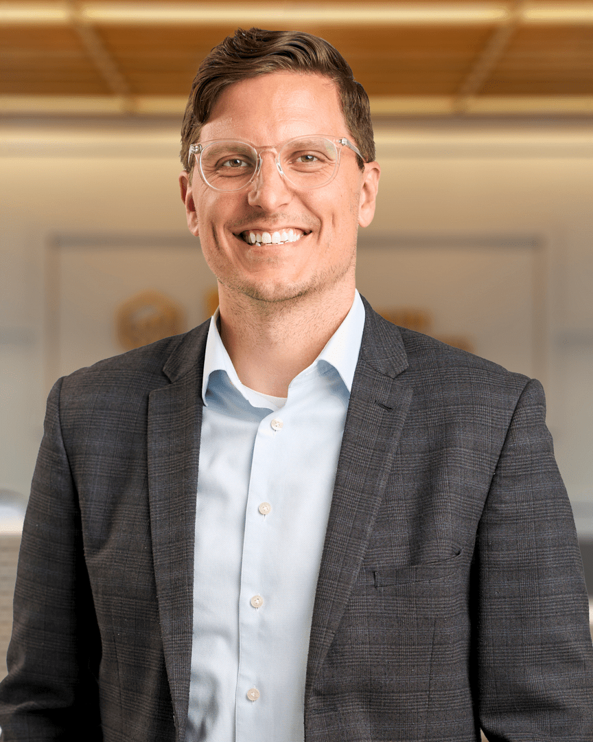 Chad, a man with short brown hair and clear framed glasses smiling in a dark plaid blazer over a light blue shirt, professional headshot with a warm lit office background.