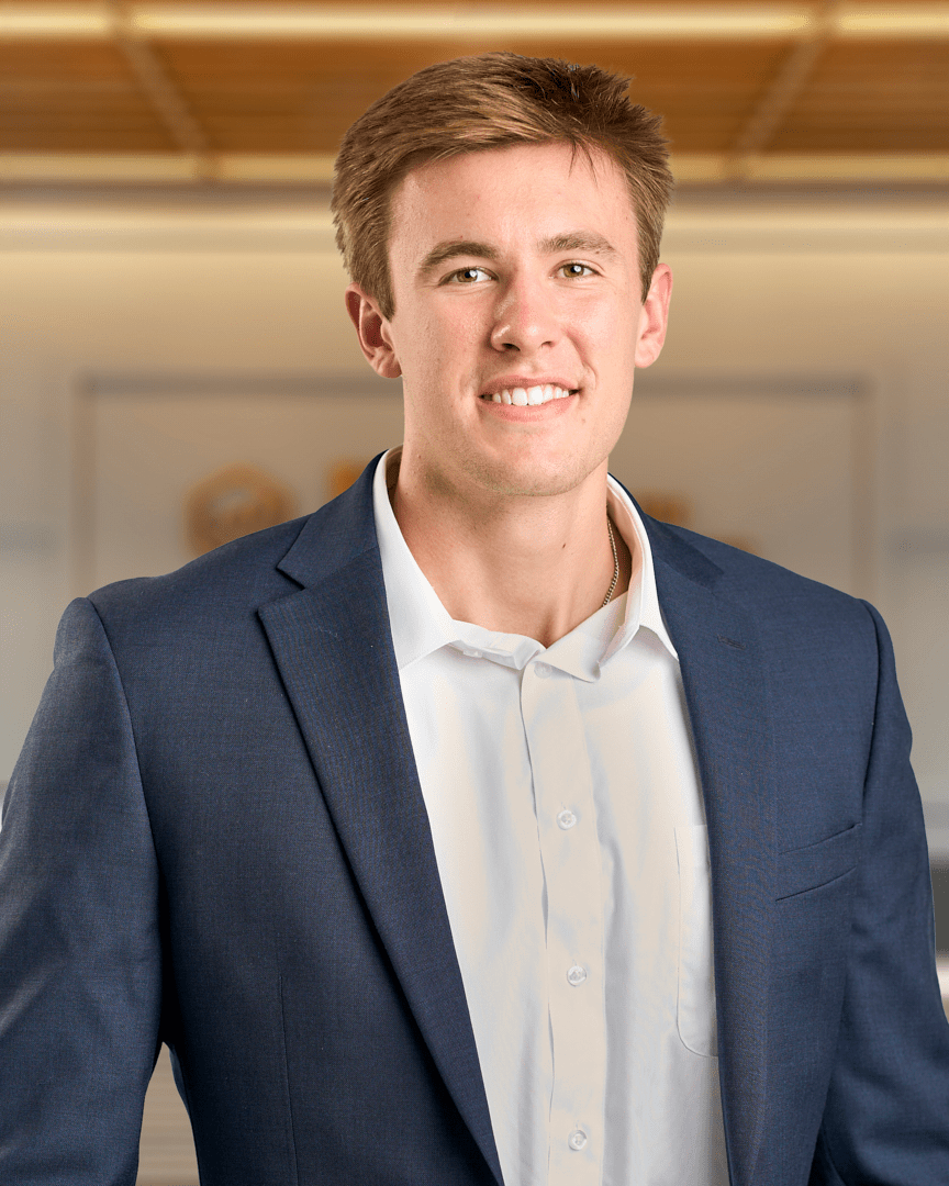 Gabe, a young man with short brown hair smiling in a navy blazer over a white button up shirt, professional headshot with a warm lit office background.