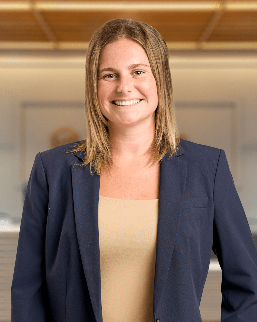Haley, a woman with shoulder length light brown hair smiling in a navy blazer over a tan top, professional headshot with a warm lit office background.