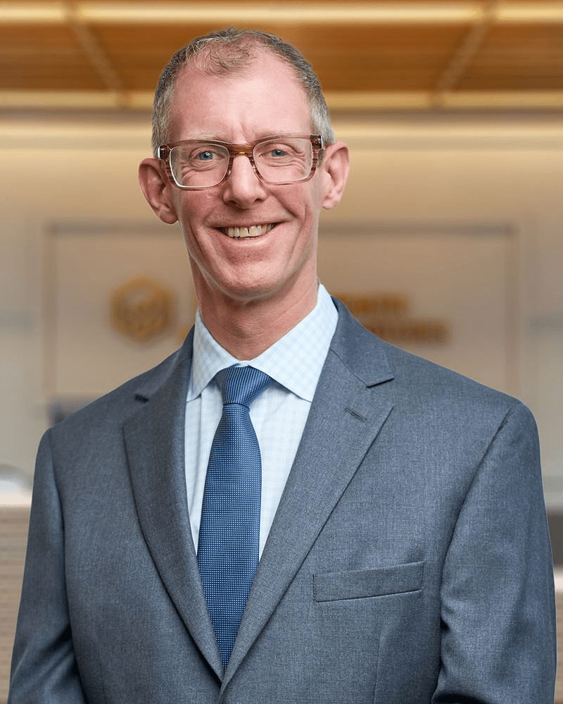 Professional headshot of a man with short gray hair, glasses, and a friendly smile, wearing a gray suit jacket, light blue checkered dress shirt, and blue textured tie, standing indoors against a blurred office background with wood ceiling accents and a gold circular logo visible behind him.