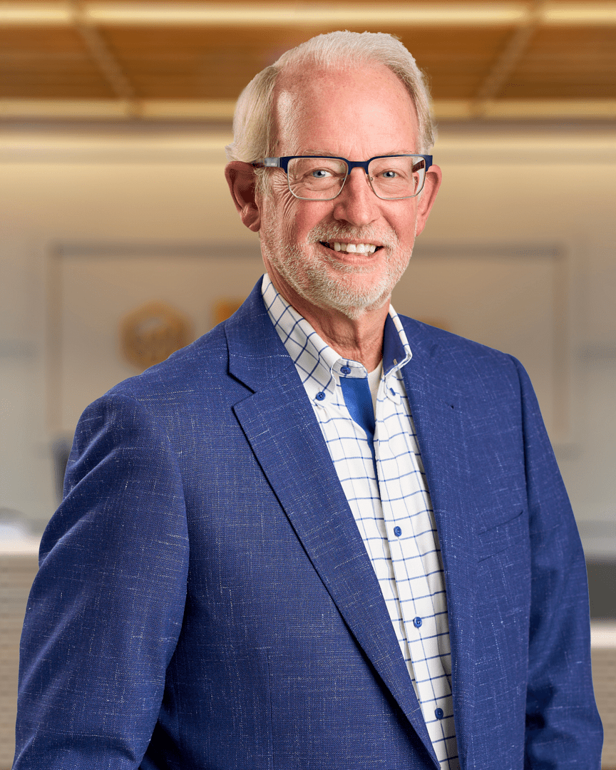 Jerry, an older man with white hair, glasses, and a short beard smiling in a blue textured blazer over a checked shirt, professional headshot with a warm lit office background.