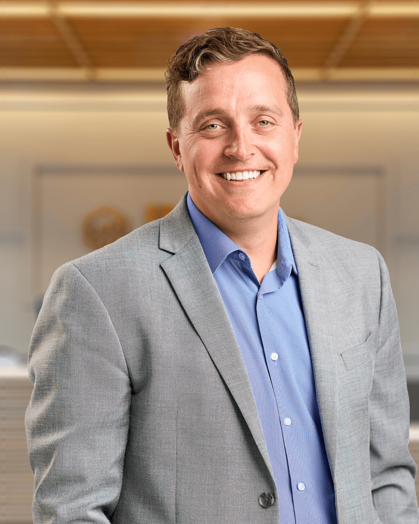 Matt K., a man with short brown hair smiling in a grey blazer over a blue button up shirt, professional headshot with a warm lit office background.